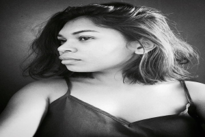 Eine selbstbewusste Frau in einem schwarzen Kleid posiert in einem Schwarz-Weiß-Foto, ihr glatter Bob und das natürliche Make-up unterstreichen ihre selbstbewusste Haltung an einer Wand.