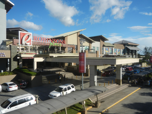 Ein belebtes Einkaufszentrum mit verschiedenen Gebäuden, Geschäften, Fahrzeugen auf der Straße, Fußgängern auf dem Gehweg, Straßenlaternen, Informationsanzeigen, Grünflächen und einer bewölkten Himmel.