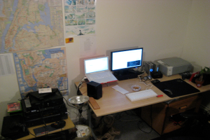 Ein Raum mit einem Tisch, auf dem ein Laptop, Lautsprecher, Monitor, Tastatur, Ball, Maus, Scanner und Papiere stehen; ein weiterer Tisch mit Elektronikgeräten in der linken unteren Ecke; und eine Wand mit Charts und Postern im Hintergrund.