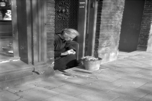 Eine ältere Frau sitzt auf einem Gehweg und hält ein Objekt in der Hand, daneben steht ein Eimer, links ist ein Schaufenster zu sehen und im Hintergrund eine Wand mit einer Tür.