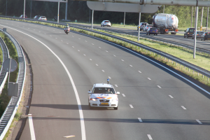Ein Auto fährt auf einer Autobahn mit einem Polizeiwagen am Straßenrand, anderen Fahrzeugen, Leitplanken, Pfosten, Schildern und Bäumen im Hintergrund.