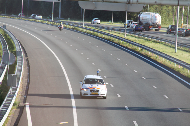 Ein Auto fährt auf einer Autobahn mit einem Polizeiwagen am Straßenrand, anderen Fahrzeugen, Leitplanken, Pfosten, Schildern und Bäumen im Hintergrund.