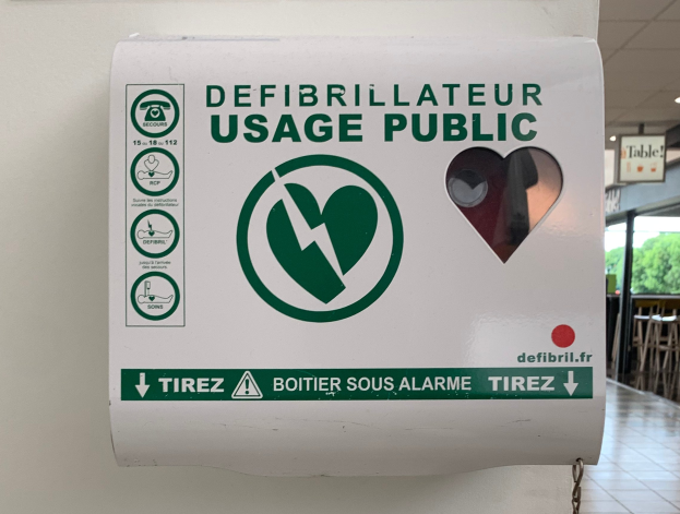 Öffentliches Schild zur Defibrillatornutzung an einer Gebäudewand angebracht, mit Stühlen, Tischen, Plakaten, Lampen und Bäumen im Hintergrund und dem Boden unten sichtbar.