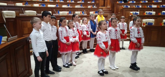 Kinder in weißen und roten Kleidern stehen in einem Raum mit einem Podium, Mikrofon, Stühlen, Tischen und Gegenständen im Hintergrund, tragend Flaggen als Teil einer Nationalversammlung.