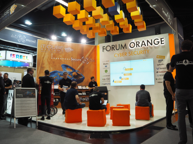 Eine Gruppe von Menschen an einem Messestand für IT-Cube Systems Forum Orange Cyber Security, mit sitzenden und stehenden Besuchern, einem Anzeigebildschirm, Regalen mit Gegenständen, Texttafeln und Deckenleuchten.