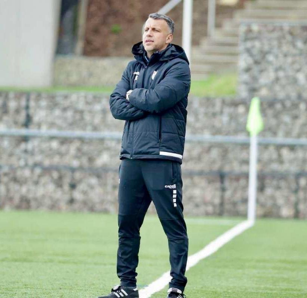 Ein Fussballtrainer mit verschränkten Armen auf einem Feld stehend, mit einer schwarzen Jacke und Hose bekleidet, mit einem Zaun, Treppe und Wand im Hintergrund.
