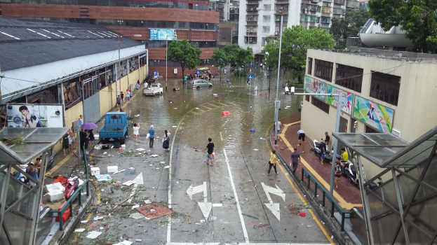 Eine überflutete Stadtstraße mit Menschen, einige halten Schirme, Gebäude, Bäume, Laternenpfähle, Fahrzeuge, verstreute Papiere und ein Schuppen auf der rechten Seite.