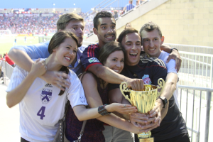 Sechs Menschen, darunter zwei Frauen und vier Männer, halten einen Pokal und lächeln für ein Foto vor einem Eisengeländer, mit einer Wand und einem Geländer oben rechts und Menschen und Lichtern im Hintergrund, möglicherweise in einem Stadion.