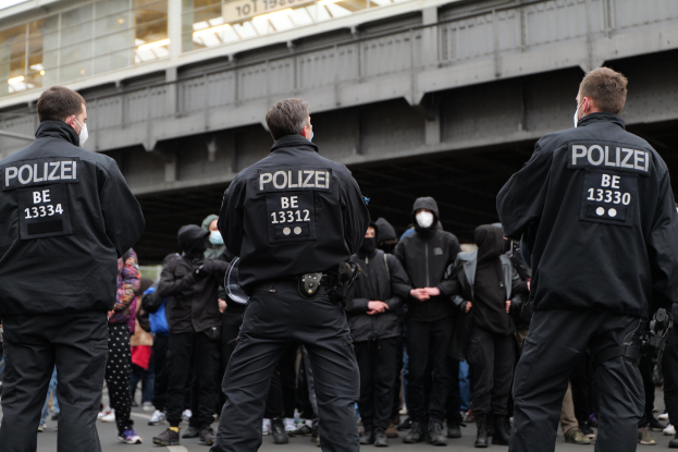 Polizisten in schwarzen Uniformen und Masken vor einer Menge mit einer Brücke und einem Gebäude im Hintergrund während einer Demonstration.