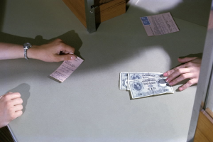 Eine Person gibt Geld an eine andere an einer Kasse ab, mit Geldscheinen und Papieren auf dem Tisch und einer Tür rechts.