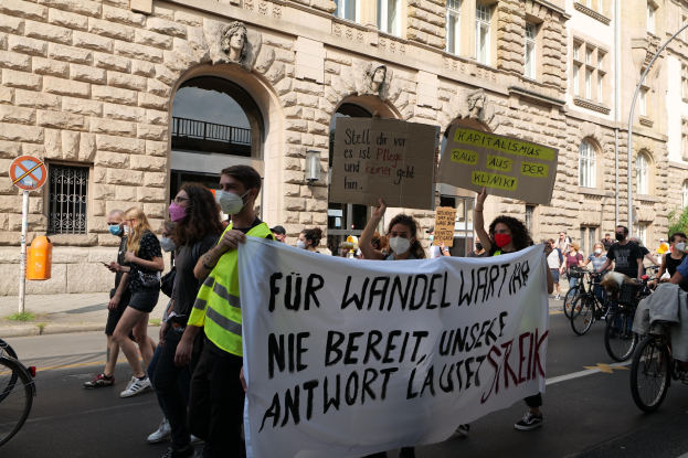 Eine Gruppe von Menschen, die auf einer Straße während einer Demonstration in Berlin gehen, einige halten Schilder und Banner, andere fahren Fahrräder, mit einem Gebäude im Hintergrund, das Fenster, Bögen, Säulen und Skulpturen hat.