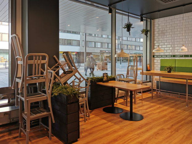 Ein Restaurant-Innenraum mit Holztischen und -stühlen, großen Fenstern, die eine Berliner Straßenszene mit Passanten, Gebäuden und Straßenelementen zeigen.