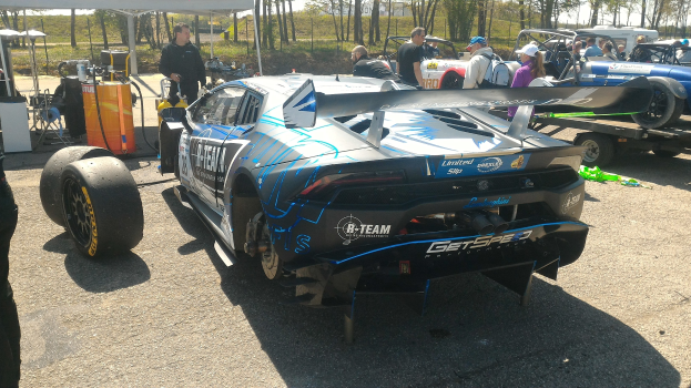 Ein Lamborghini Huracan GT3 Rennauto auf einem Parkplatz mit Menschen in bunten Kleidern, einem Zelt links daneben, Bäumen und einem klaren blauen Himmel im Hintergrund.