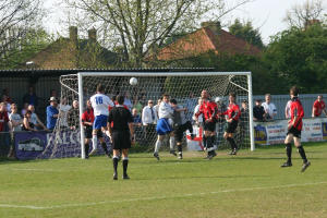 Players are playing football on a field with a goal net behind them, while spectators stand and watch from the sidelines, with trees and houses visible in the background.