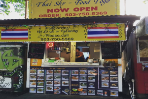 Ein Geschäft mit verschiedenen Artikeln wie Getränkedosen, Speisekarten, einer Uhr, Gewürzen in Plastikbehältern, Gewürzen, Werbetafeln und Namensschildern unter einem Himmel.