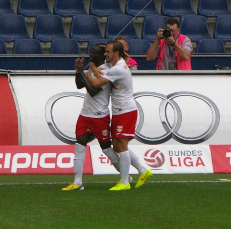Zwei Fussballspieler feiern ein Tor auf einem Feld mit einer Menge, einem Fotografen, einer Fahne, Stühlen und einer Wand im Hintergrund.