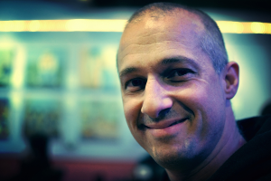 Ein Mann in einem schwarzen Kleid lacht mit einem unscharfen, beleuchteten Hintergrund.