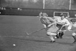 Schwarzes Bild einer Gruppe von Frauen, die Feldhockey auf einem Feld spielen, mit einem Ball im Spiel, einem Torpfosten und Zuschauern im Hintergrund.