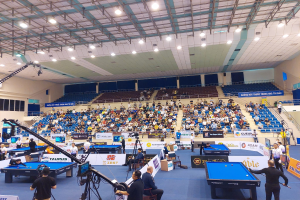 Ein großer Raum mit mehreren Billardtischen, umgeben von sitzenden Spielern, Fenstern an der linken Wand, Deckenbeleuchtung und einer Kameraaufstellung, die auf ein professionelles Pool-Turnier hinweist.