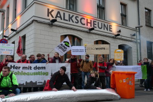 Eine Gruppe von Menschen hält Schilder und Plakate in der Hand und steht vor einem Gebäude, wobei zwei Personen im Vordergrund und ein Müllcontainer rechts zu sehen sind, während sie in Deutschland protestieren.