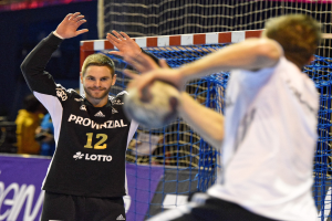Zwei Männer spielen Handball vor einem Tor mit Netz und Banner im Hintergrund in einer Stadionatmosphäre.
