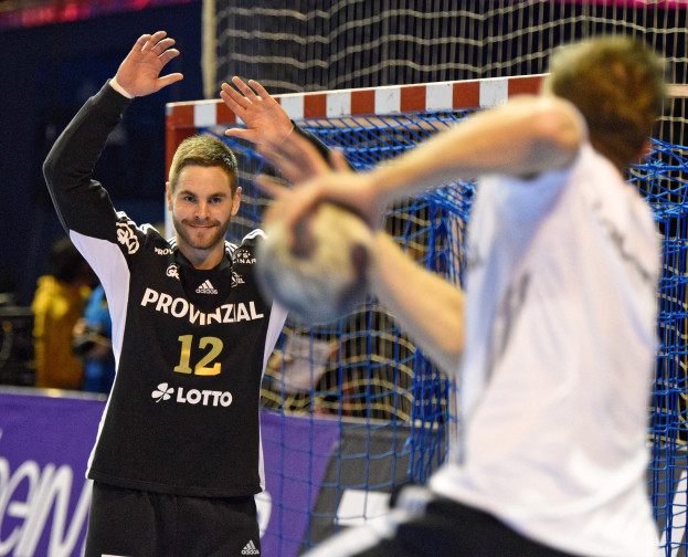 Zwei Männer spielen Handball vor einem Tor mit Netz und Banner im Hintergrund in einer Stadionatmosphäre.