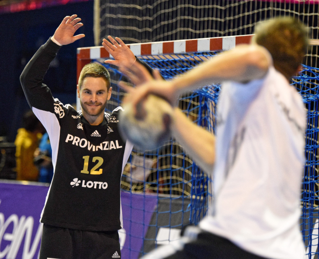 Zwei Männer spielen Handball vor einem Tor mit Netz und Banner im Hintergrund in einer Stadionatmosphäre.