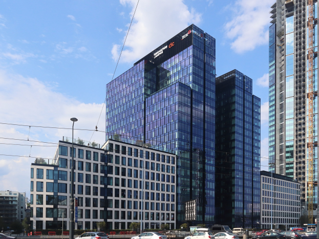 Großes modernes Bürogebäude mit vielen Fenstern, identifiziert als die Deutsche Bank Hauptstad in Frankfurt, Deutschland, umgeben von städtischer Infrastruktur einschließlich Straßenlaternen, Fahrzeugen, Bäumen und einem bewölkten Himmel.