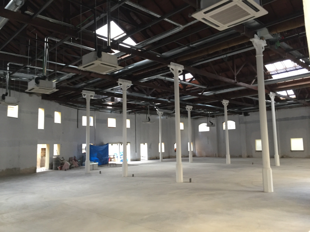 Große Industriehalle mit zahlreichen Fenstern, tragenden Säulen, einer hohen Decke mit Beleuchtung und Klimaanlagen sowie einem Betonboden und -wänden.