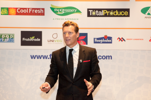 Ein Mann in einer schwarzen Blazer steht mit einem Gegenstand in der Hand und spricht, vor einem weißen Banner und einer weißen Tafel mit Organisationslogos im Hintergrund.