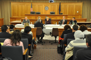 Ein Sitzungssaal mit einer Gruppe von Menschen, die auf Stühlen sitzen, zwei Flaggen anderer Länder, ein oranger Schreibtisch mit Wasserflaschen und einem Mikrofon.
