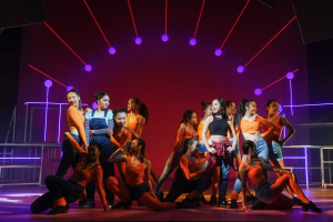 Eine Gruppe junger Tänzer in bunten Kostümen performs auf einer Bühne mit Geländern, die Arme erhoben, während das Publikum unter bunten Lichtern und gemusterten Bühnendekorationen klatscht und jubelt.