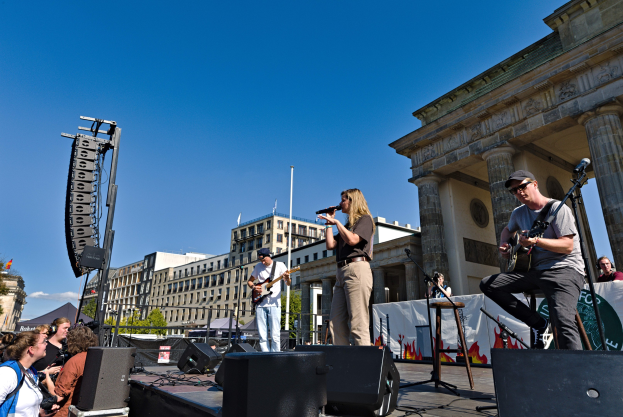 Eine Gruppe von Menschen, die auf einer Bühne vor dem Brandenburger Tor Musik spielen, mit Lautsprechern und Equipment drumherum, unter einem klaren blauen Himmel.