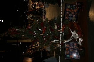 Ein Weihnachtsbaum mit Geschenken, Rahmen und Spielzeugen auf einer Plattform, mit Menschen, zusätzlichen Weihnachtsbäumen, einer Kette, einem Pfahl und Lichtern im Hintergrund.