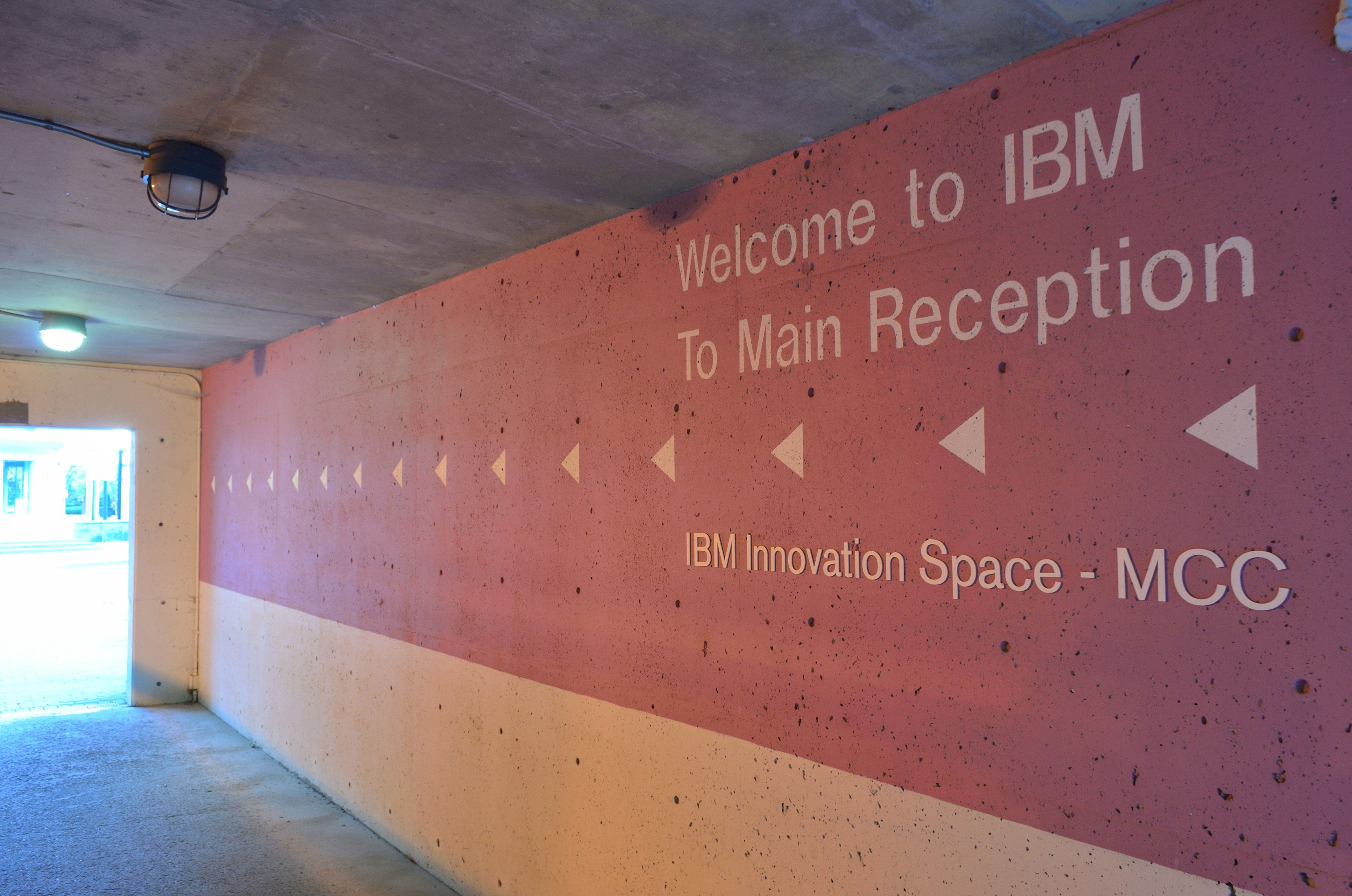 Eine Wand mit "Willkommen bei IBM" beleuchtet von Deckenlampen und eine Tür im Hintergrund mit der Aufschrift "IBM Innovationsraum MCC."