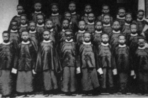 Ein Schwarz-Weiß-Foto einer Gruppe von Kindern, die vor einem Gebäude mit einer Wand und einer Tür im Hintergrund stehen, wahrscheinlich eine afroamerikanische Schule.