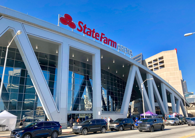 Außenansicht der State Farm Arena in Minneapolis, Minnesota, mit Glaswänden, Säulen, einem Namensschild oben, Laternen, Fahrzeugen, Fußgängern, Bäumen und einem klaren blauen Himmel.