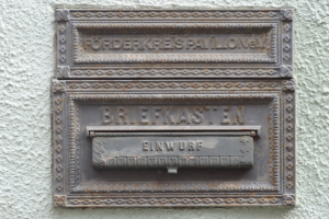 Alter rechteckiger Metallbriefkasten mit einem oberen Griff, an einem Gebäude montiert, beschriftet mit "Briefkasten" in schwarzer Schrift.