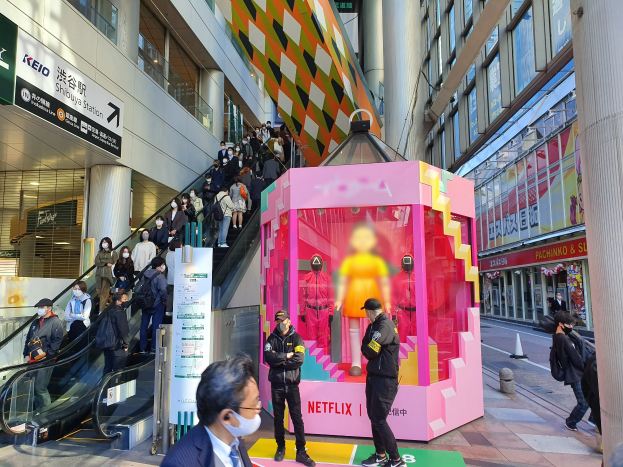 Eine Gruppe von Menschen steht vor einem Gebäude mit Glaswänden, Säulen und Texttafeln, einige tragen Masken, mit einer Rolltreppe im Blick; eine pinkfarbene Box in der Mitte zeigt Schaufensterpuppen und trägt die Aufschrift "Netflix' neue Netflix-Bühne in Tokio."
