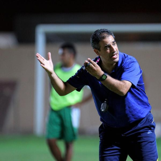 Ein Mann in einem blauen T-Shirt und Hose steht auf einem Fußballfeld und hält ein Mikrofon in der Hand, während er gestikuliert. Im Hintergrund ist eine Person in einem grünen Kleid sowie ein Torpfosten und eine Wand zu sehen.