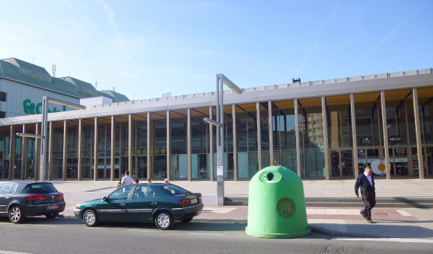 Grüne Mülltonne neben der Straße in der Nähe des Berliner Hauptbahnhofs, die Glaswände und Säulen hat, mit Fahrzeugen und Passanten in der Nähe unter einem bewölkten Himmel.