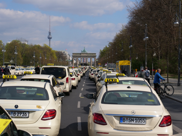 Lange Reihe von Taxis, die an einer belebten Straße in Berlin, Deutschland, geparkt sind, mit Fahrradfahrern und Fußgängern auf dem Bürgersteig, flankiert von Bäumen und Laternen, und Gebäuden, einem Bogen und einem Turm im Hintergrund unter einem bewölkten Himmel.