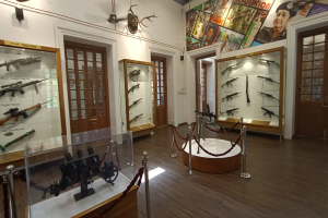 Ein Raum voller verschiedener Waffenarten, darunter eine Maschinenpistole in einer Glasvitrine auf einem Tisch, mit einem Seilzaun, Pfosten, einem Banner mit Bildern und Text an der Wand, Türen und Deckenlampen, die zu einem Dach führen.