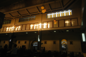 Das Innere einer Kirche mit Menschen auf Bänken sitzend und auf dem Boden stehend, Wände mit Fenstern geschmückt und von der Decke hängende Lampen.