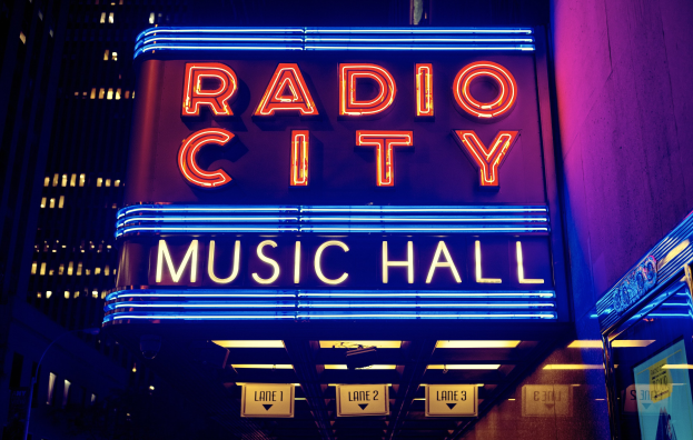Außenansicht des Radio City Music Hall in New York City bei Nacht, hell erleuchtet mit hohen Gebäuden im Hintergrund, mit einer 'Radio City'-Leuchtschrift und mehreren kleineren Leuchtschriften auf der rechten Seite.