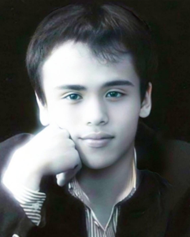 Ein selbstbewusster junger Mann in einer schwarzen Jacke posiert vor einem dunklen Hintergrund mit entschlossener Miene.