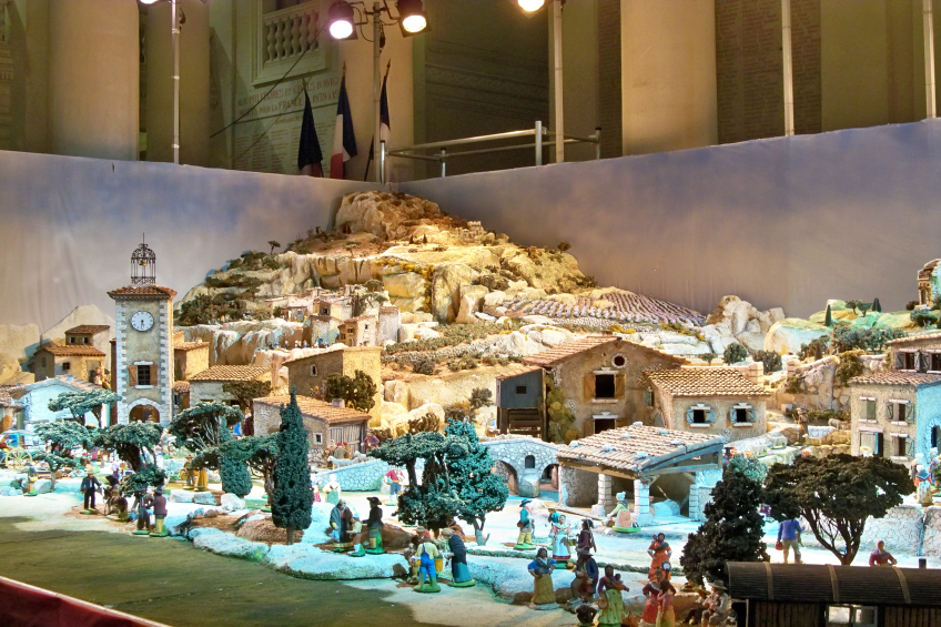 Eine Museumsausstellung eines Krippenmodells mit Menschen, Häusern, Bäumen, Steinen, einem Kirchturm, Flaggen, Pfählen und Lichtern, alles beleuchtet von Museumsbeleuchtung.