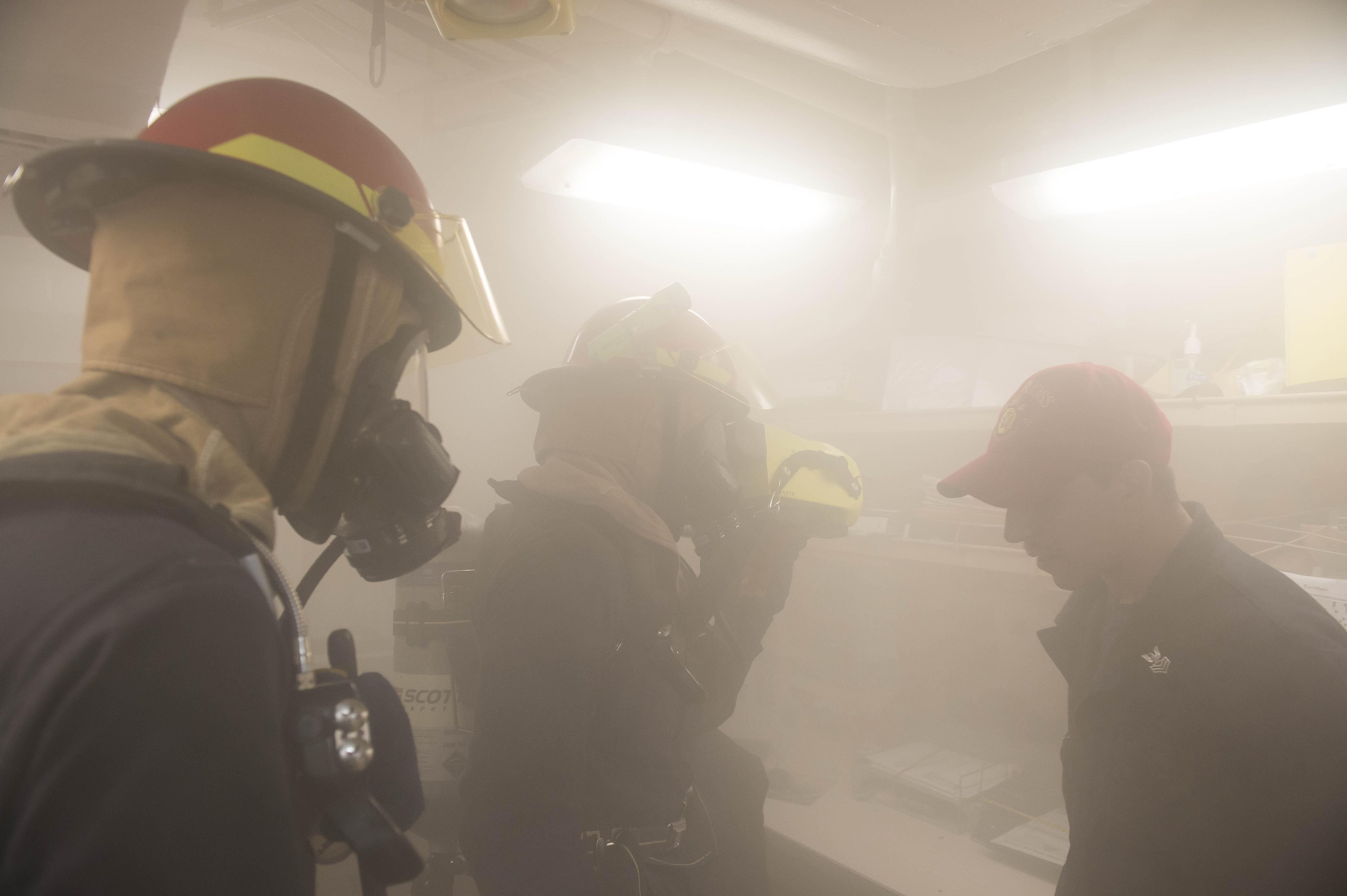 Feuerwehrleute in einem rauchgefüllten Raum während einer Übung, einer hält eine Kamera, trägt Helme mit Rauch aus dem Mund.