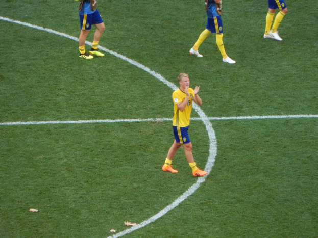 Eine Gruppe von Männern in Fußballtrikots und Stollenschuhen steht auf einem Fußballfeld, wobei eine Person in der Mitte klatscht.