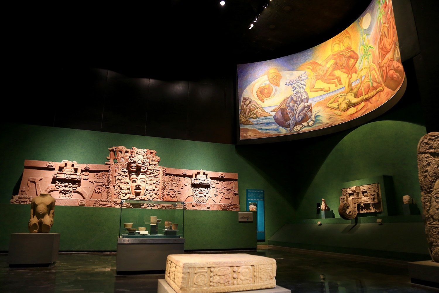 Ein Museumsraum mit verschiedenen Artefakten, darunter Skulpturen, Tischgegenstände, eine Texttafel, eine Wandmalerei und Deckenbeleuchtung im Anthropologischen Museum von Mexiko-Stadt.
